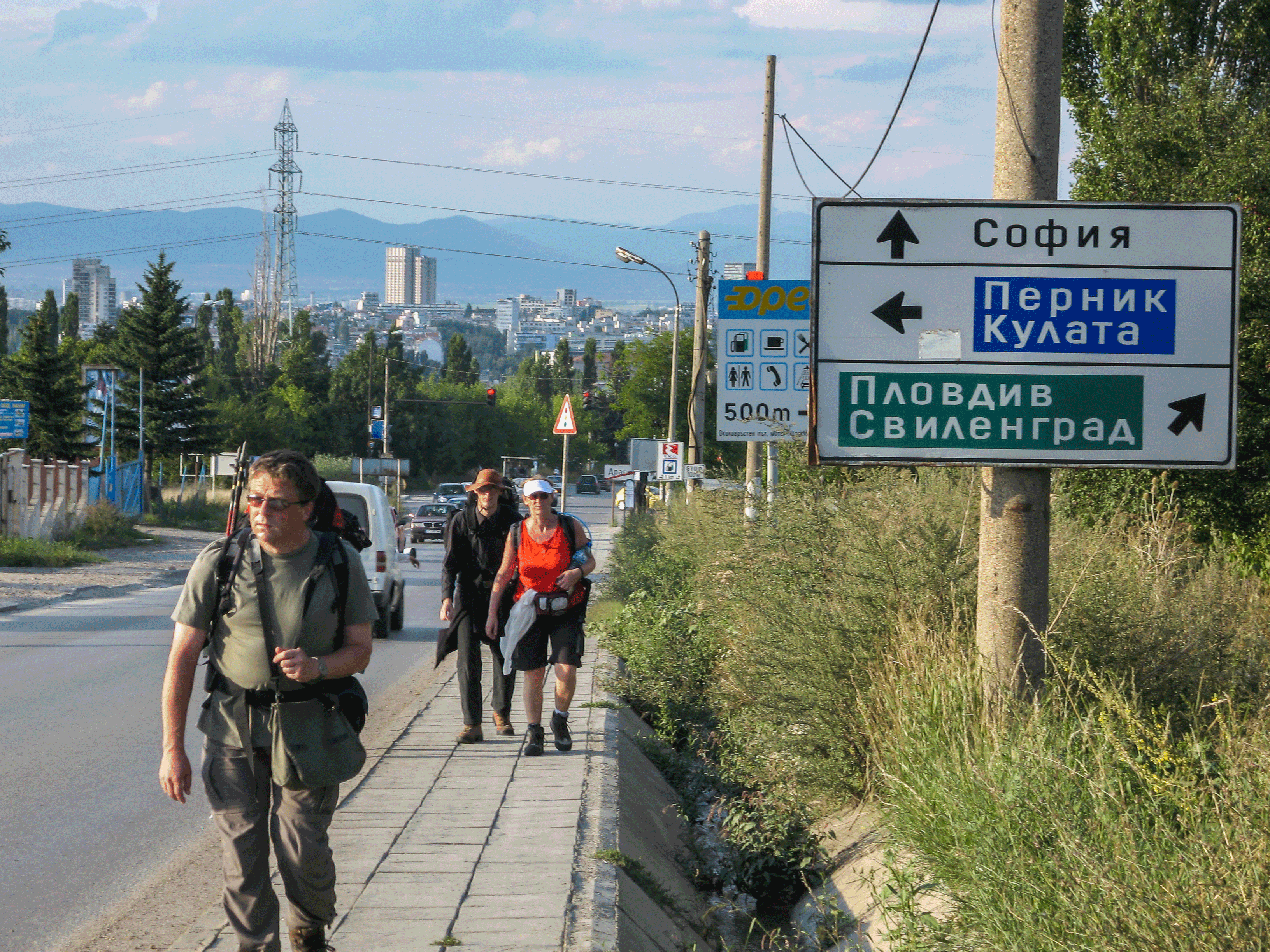 Wie das Straßenschild beweist: Hinter uns liegt Sofia, links geht‘s zum Endziel des E4 in Bulagiern: Kulata Wanderer auf einem Bürgersteig entlang einer Straße, umgeben von einer Stadtlandschaft mit Gebäuden und Bergen; rechts ein Straßenschild mit Wegweisern nach Sofia, Pernik/Kulata, Plovdiv und Svilengrad