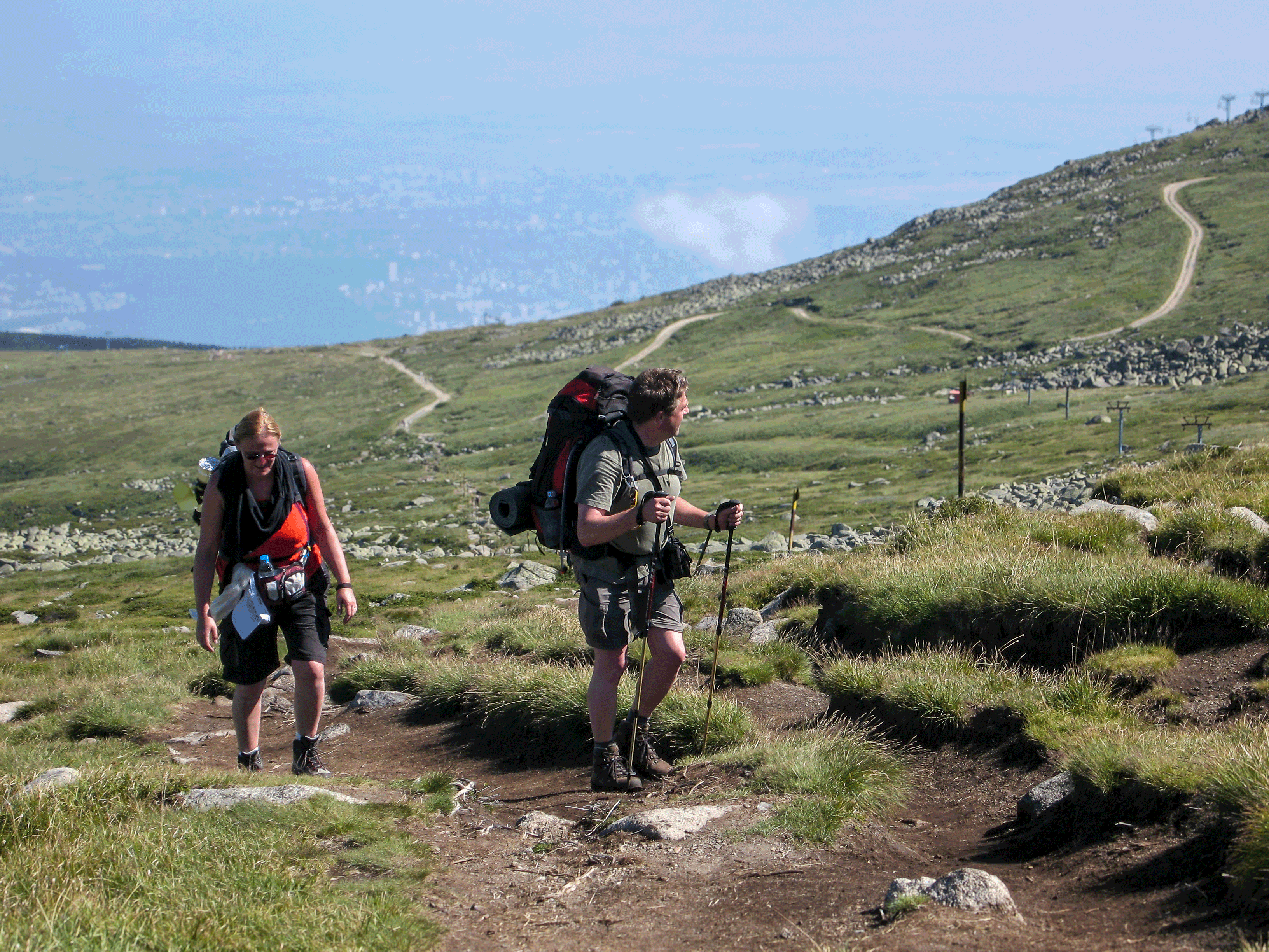 Die höchste Erhebung der ersten Tour, der Cernie Vrah, ist gleich geschafft. Im Hintergrund lässt sich die bulgarische Hauptstadt zu den Füßen des Vitoscha- Gebirges erahnen Wanderer auf einem felsigen Pfad durch grüne Hügel mit blauem Himmel im Hintergrund – Natur, Abenteuer, Wandern in bergiger Landschaft.