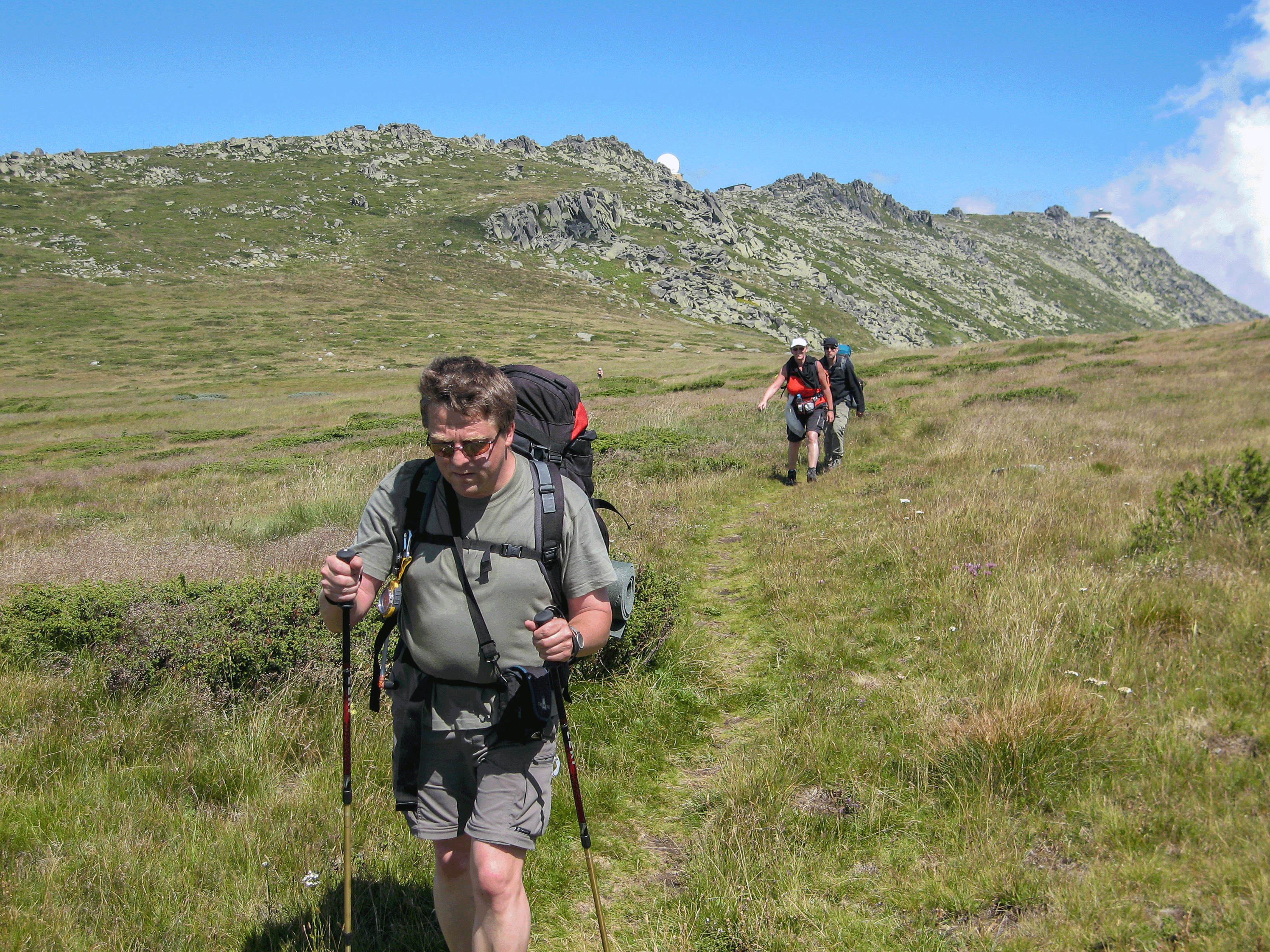 Gut zum Einwandern: ein herrlicher Weg. Im Hintergrund guckt nicht etwa der Mond um die Ecke - es ist die Kugel der Wetterstation Drei Wanderer gehen einen Pfad durch eine hügelige Landschaft aus Gras und Felsen entlang. Der Himmel ist klar und blau, und der Wanderer im Vordergrund trägt einen großen Rucksack und verwendet Trekkingstöcke.