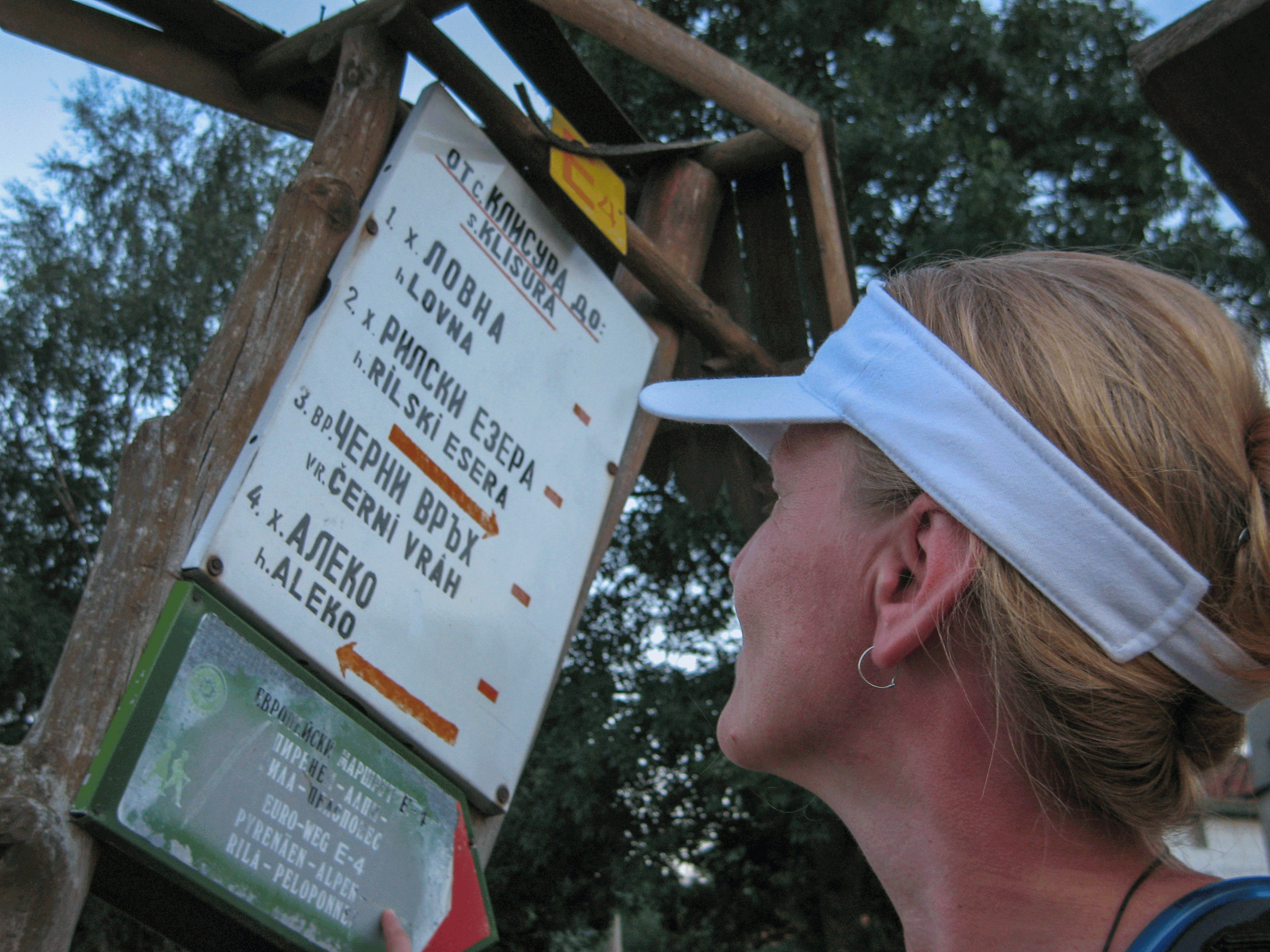 Solch feine Schilder, noch dazu in lateinischer und kyrillischer Schrift sind selten auf unserer Tour Eine Wanderin betrachtet ein hölzernes Wegweiser-Schild mit Wanderzielen in kyrillischer Schrift, einschließlich Hinweise auf den Europäischen Fernwanderweg E 4.