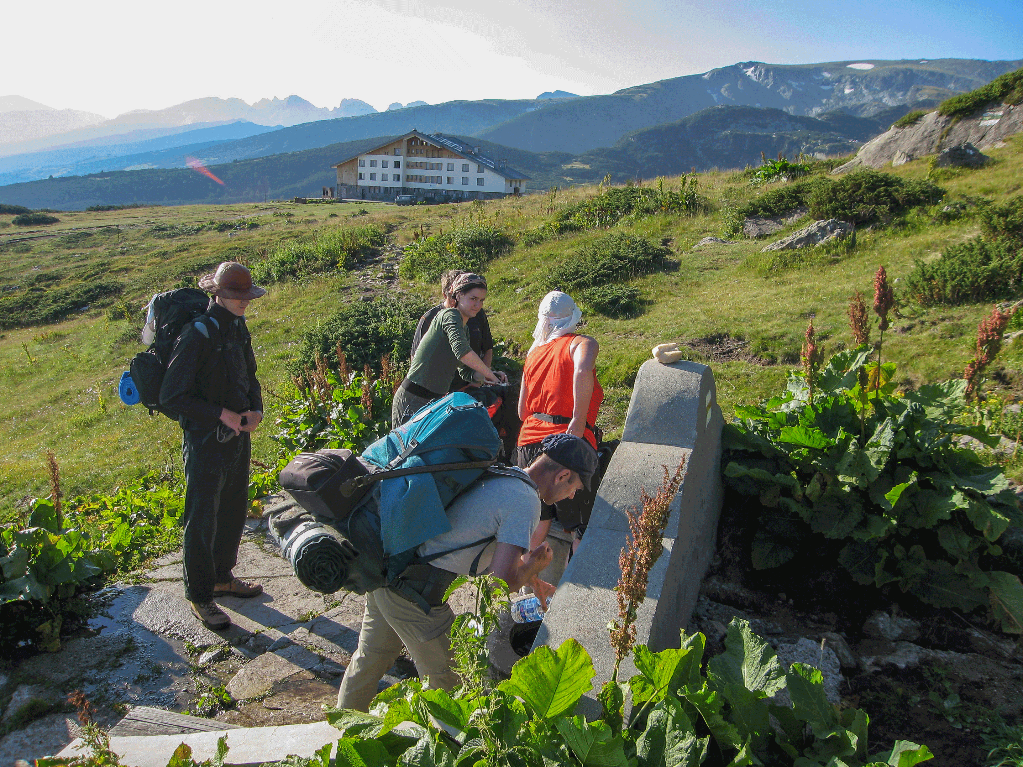 An der Quelle werden die Wasservorräte aufge-frischt. Im Hintergrund eine der modernsten Hütten Bulgariens: die Rilski Ezera Wanderer befüllen Wasserflaschen an einem alpinen Brunnen, mit Berghaus und grüner Landschaft im Hintergrund.