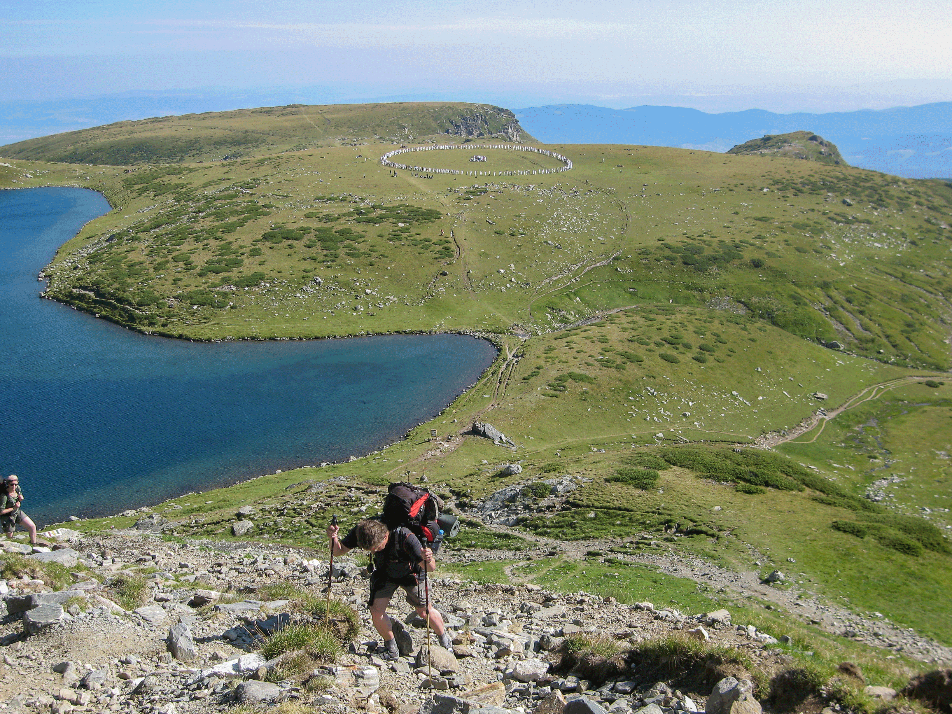 Dieses Plateau, auf dem jetzt die „White brothers“ im Kreis tanzen, ist nun überquert - jetzt geht‘s richtig in den Berg Ein Wanderer erklimmt mit Rucksack und Wanderstöcken einen steinigen Hang in alpiner Landschaft, mit einem blauen Bergsee und grünen Hügeln im Hintergrund.